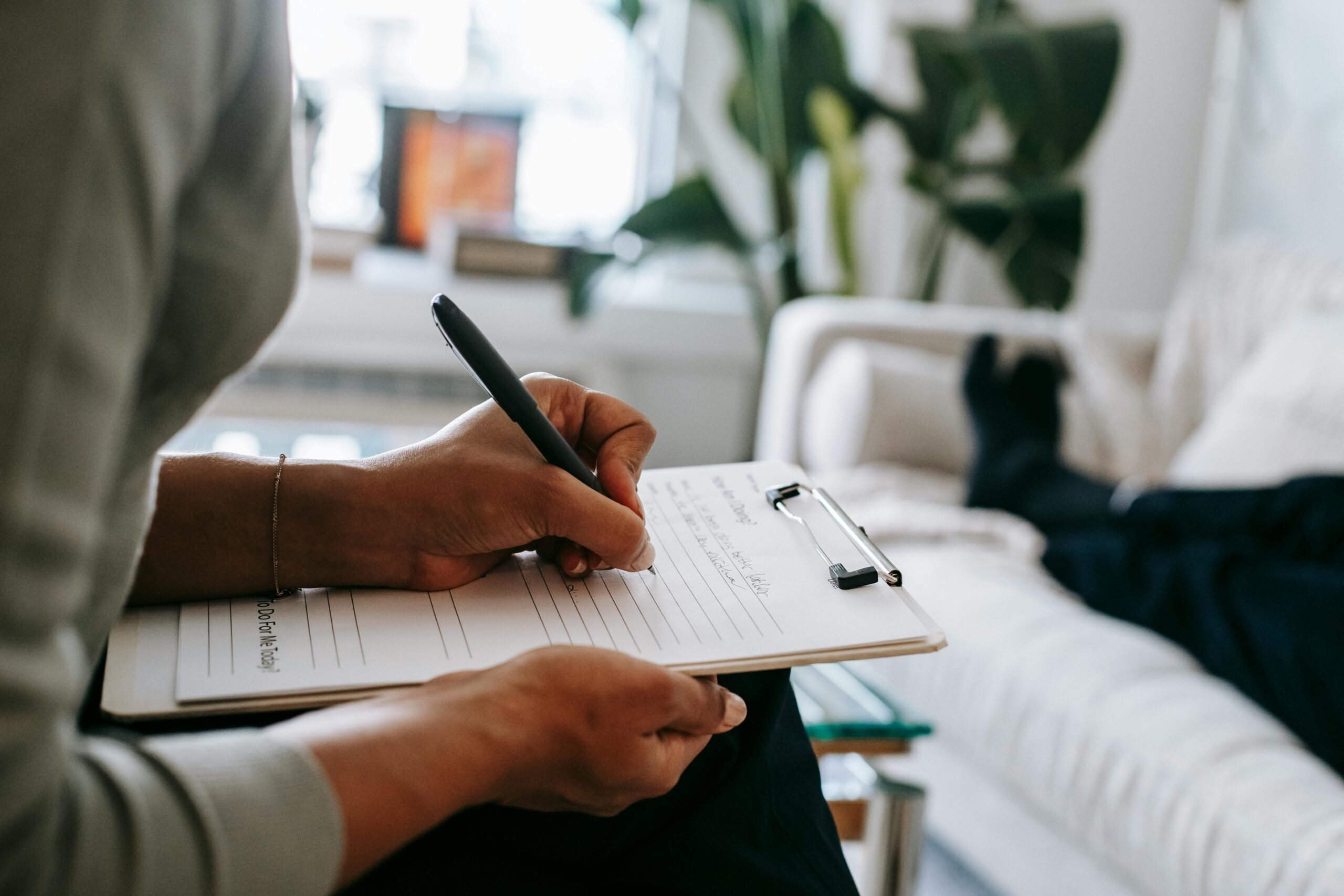 Unrecognizable ethnic female therapist taking notes on clipboard while filling out form during psychological appointment with anonymous client lying on blurred background